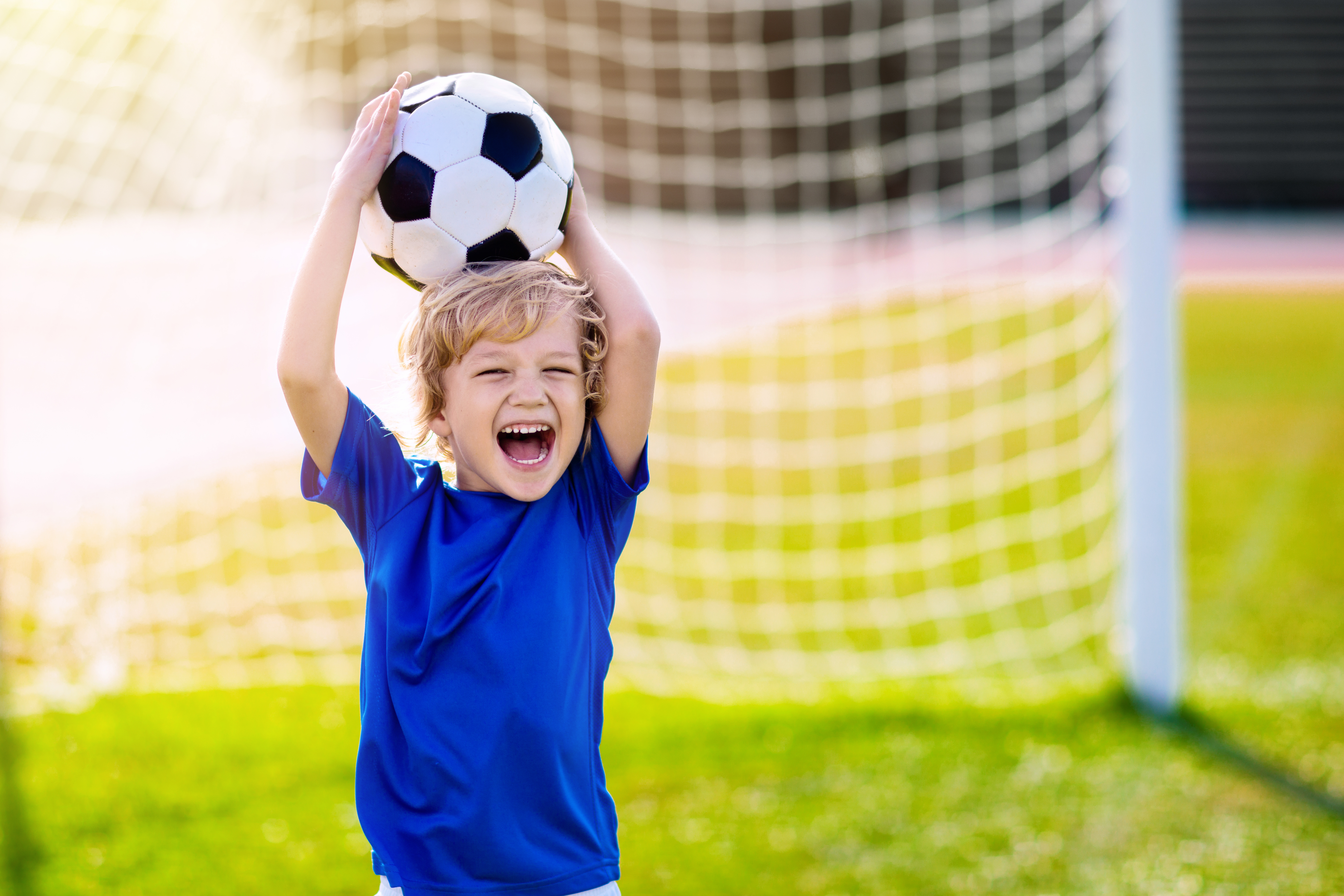 young boy holding a soccer ball over his head smiling with a soccer net behind him on a grass field