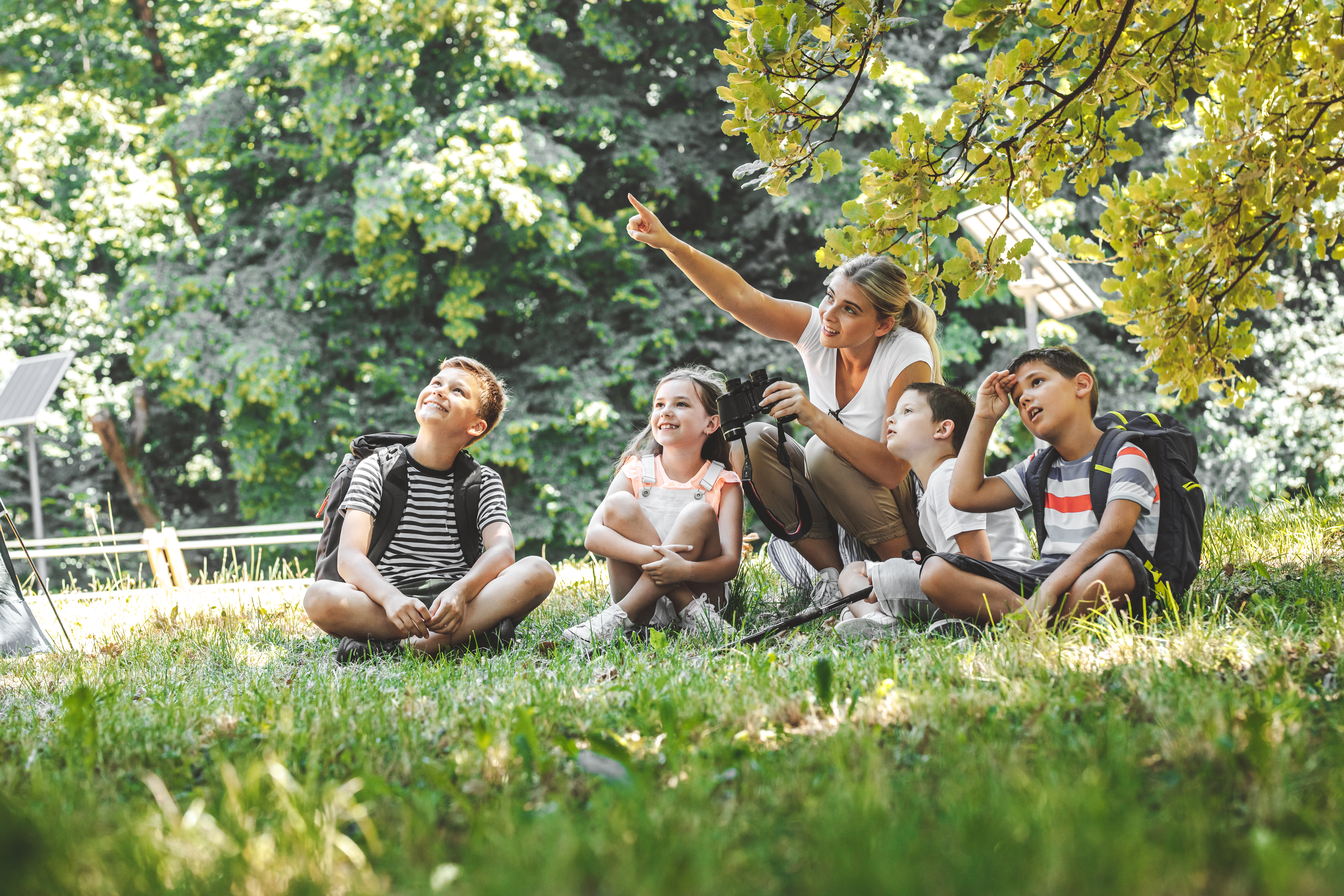 kids sitting in a circle in a grass field looking at something the camp counselor is point at